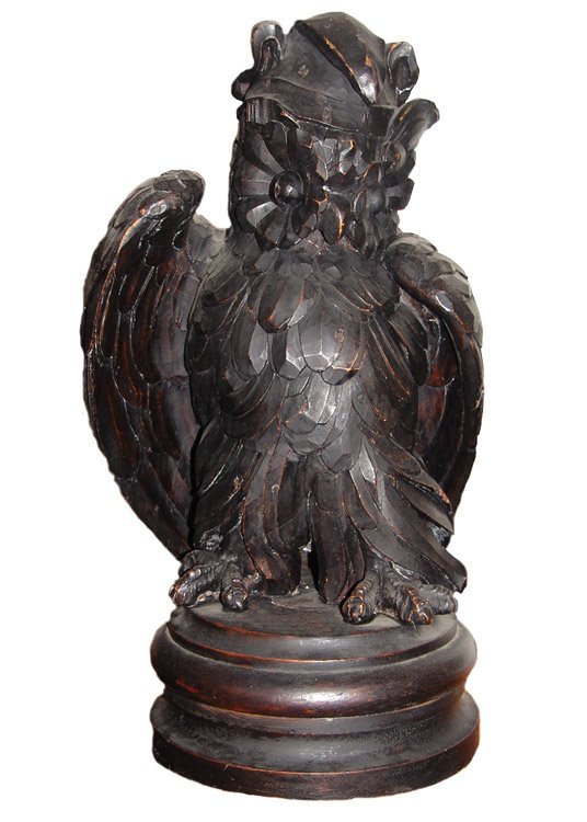 Der geschnitzte Praga-Uhu aus dunklem Holz, gefiedert herausgearbeitet, Flügel angelegt, auf einem gedrechselten Sockel, freigestellt vor hellem Grund